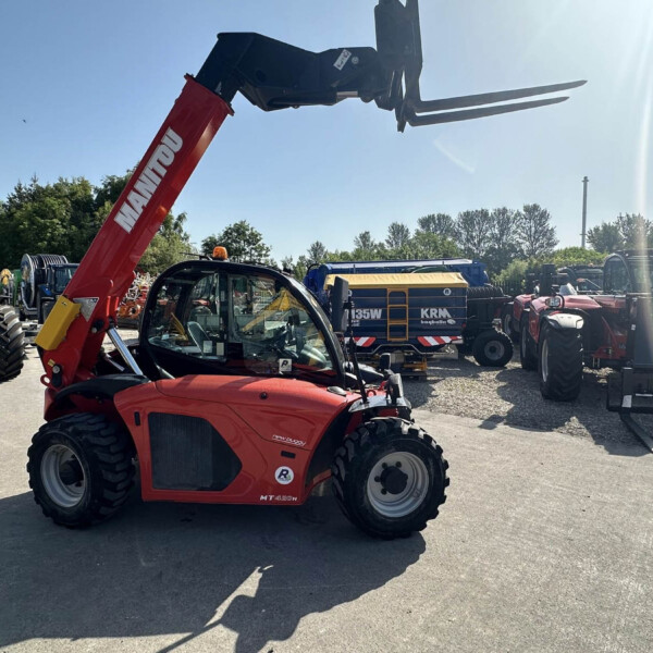Red Manitou telescopic handler with forks raised, in a yard full of tractors and farm machinery under a clear blue sky.