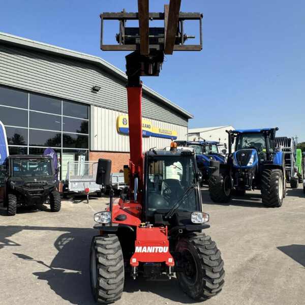 Red Manitou forklift with its forks raised high in an outdoor equipment yard, with blue tractors and a building in the background.