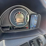 Dashboard cluster with analog RPM gauge and a rectangular digital display showing vehicle data, surrounded by control buttons on the steering console panel.