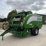 Green agricultural baler parked outside a gray industrial building, with a tractor wheel visible on the right and a cloudy sky above.