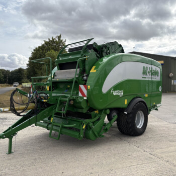 Green agricultural baler parked outside a gray industrial building, with a tractor wheel visible on the right and a cloudy sky above.
