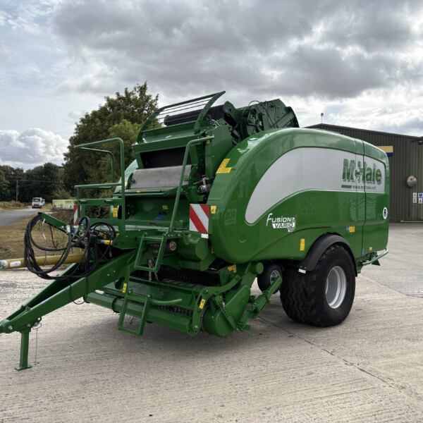 Green agricultural baler parked outside a gray industrial building, with a tractor wheel visible on the right and a cloudy sky above.