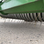 Close-up of a green agricultural harvester header with curved metal tines near the ground under the machine frame.