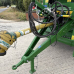 Close-up of green agricultural machine with tangled hydraulic hoses and a yellow beam on a gravel road, outdoors.