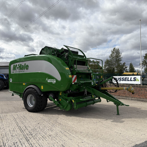 Green McHale Fusion baler on a concrete lot with a blue tractor nearby and a cloudy sky above, ready for field work