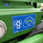 Close-up of a green industrial control panel with a round pressure gauge and a blue instructional sticker on the surface, hoses running nearby.