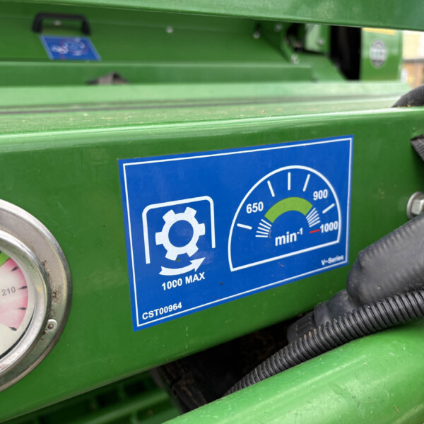 Close-up of a green industrial control panel with a round pressure gauge and a blue instructional sticker on the surface, hoses running nearby.