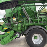 Close-up of a green agricultural harvester's internal mechanism with gears, hoses, and metal framework near a large wheel.