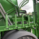 Close-up of green agricultural machinery showing hydraulic rods, hoses, and a wide black conveyor belt in a busy frame.