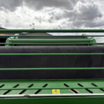 Close-up of green agricultural machinery with cylindrical rollers and a yellow warning sign, set against a cloudy sky behind.