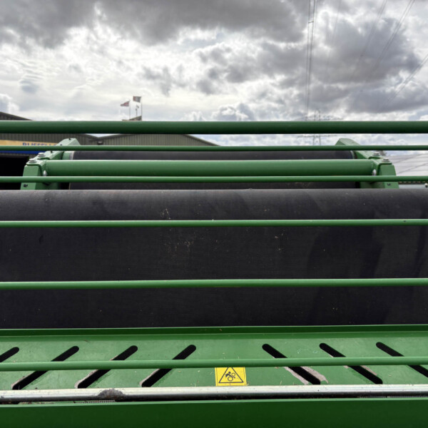 Close-up of green agricultural machinery with cylindrical rollers and a yellow warning sign, set against a cloudy sky behind.