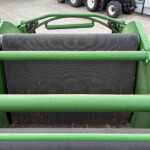 Close-up of a green farming machine with a large black conveyor roller and steel brackets.