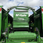 Rear view of a large green McHale Fusion VARIO baler with protective panels and warning decals, showing the machine's opening and rollers on a sunny day