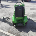 Green ride-on lawn mower with a black cushioned seat and MG logo, parked in a paved lot near a fence and cars.