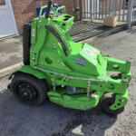 Green Mean Green riding mower parked outside a brick building with a cardboard box on the curb nearby, front wheel in view and control levers visible.