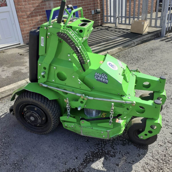 Green Mean Green riding mower parked outside a brick building with a cardboard box on the curb nearby, front wheel in view and control levers visible.