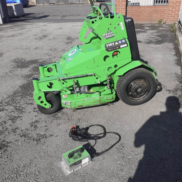 Bright green compact mower/attachment on a paved lot with cables nearby and urban buildings in background.
