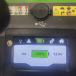 Close-up of a handheld diagnostic screen showing 98% battery, 52.9V, and USB icon with safety warning stickers above on a green and gray panel.