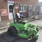 Green riding lawn mower parked outside a brick building near a reception sign.
