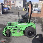 Green riding lawn mower with a black seat and tall roll bar parked in a paved lot beside a brick wall and metal fencing, with urban buildings in the background.