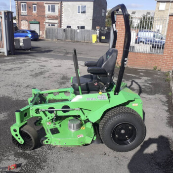 Green riding lawn mower with a black seat and tall roll bar parked in a paved lot beside a brick wall and metal fencing, with urban buildings in the background.