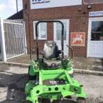 Green ride-on lawn mower facing camera, parked outside a brick building with Russell sign in the background.