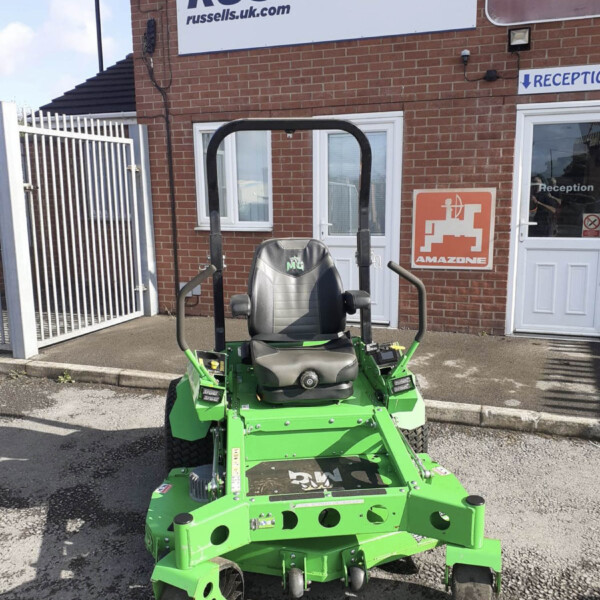 Green ride-on lawn mower facing camera, parked outside a brick building with Russell sign in the background.