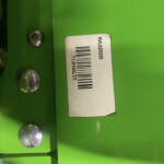 Close-up of a white barcode label on a bright green metal surface with visible screws along the edge; label includes alphanumeric text and a barcode.