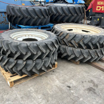 Stacks of large tractor tires on wooden pallets in an industrial yard with blue machinery nearby
