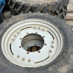 Two large tractor tires with white, rusted rims stacked on a dusty workshop floor, showing rugged treads and metal bolts.