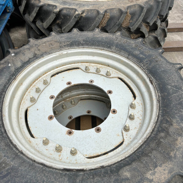 Two large tractor tires with white, rusted rims stacked on a dusty workshop floor, showing rugged treads and metal bolts.