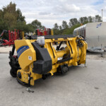 Industrial yard with bright yellow construction machine in foreground and red machinery in background, plus a silver trailer on right side under cloudy sky