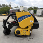 Close-up of a yellow New Holland compact loader in a gravel yard, with trailers and a green fence in the background.