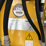 Close-up of yellow industrial machine showing hydraulic hoses, metal fittings, and safety and identification labels.
