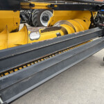 Close-up of a yellow industrial header with a large rotating roller and black rails; parts show wear and chipped paint, with a blue tractor in the background.