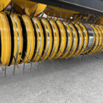 Close-up of yellow circular farming equipment with many metal tines on a gray pavement surface.