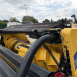 Close-up of yellow New Holland agricultural machinery with a black conveyor/guard and mesh guard in an outdoor lot.