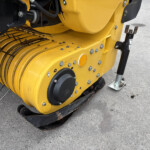 Close-up of a yellow road roller drum with metal tines and bolts on a gray concrete surface.
