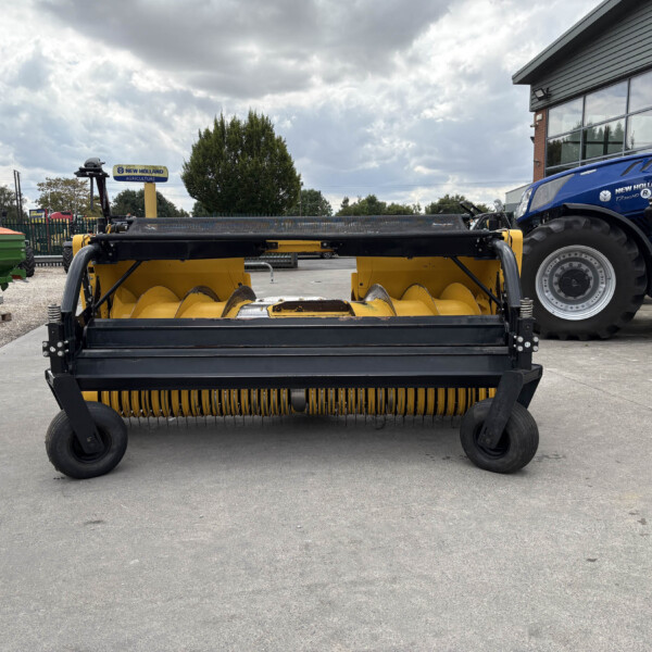 Yellow agricultural baler in the foreground with a blue New Holland tractor beside a building at a dealership facility.