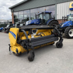 Blue New Holland tractors and a yellow harvesting implement parked outside an agricultural equipment shop at a dealership yard.