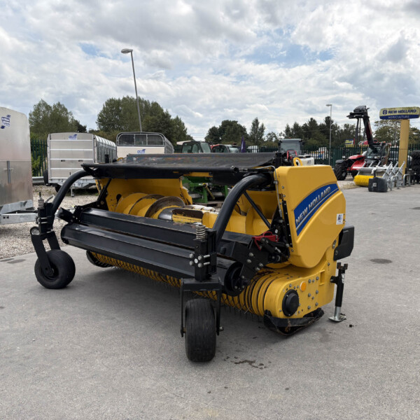 Yellow New Holland baler in a farm equipment lot with trailers and other machinery nearby.