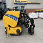 Close-up of a yellow New Holland baler in the foreground with blue tractors and a dealership building in the background.