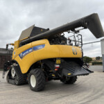 Yellow New Holland CR9.0 combine harvester with a large black unloading auger extended overhead, parked in an industrial yard under a cloudy sky.