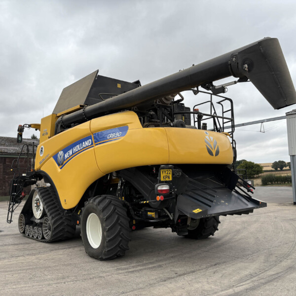 Yellow New Holland CR9.0 combine harvester with a large black unloading auger extended overhead, parked in an industrial yard under a cloudy sky.