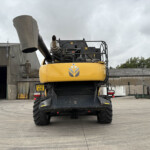 Rear view of a yellow excavator on a construction site, showing the engine housing, exhaust and platform against a cloudy sky.