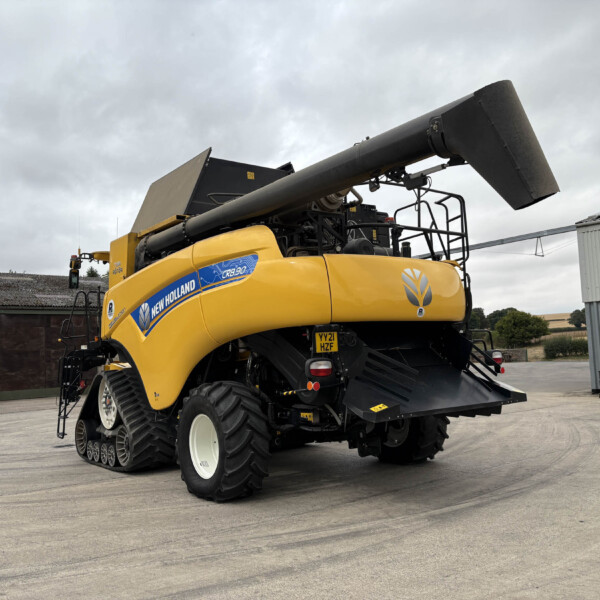 Yellow New Holland CR8.90 combine harvester with a raised grain auger in a farmyard behind cloudy skies.