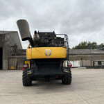 Rear view of a large yellow excavator in an industrial yard under a cloudy sky, showing its black undercarriage and machinery.