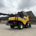 Yellow New Holland CR8.90 combine harvester with extended unloading auger in an industrial yard under cloudy skies