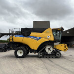 Large yellow New Holland CR8.90 combine harvester parked in an industrial yard under a cloudy sky, with a building in the background.