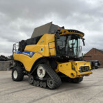 Yellow New Holland harvest combine with tracked undercarriage, parked in an industrial lot with cloudy sky in the background, front-right view, cab visible.